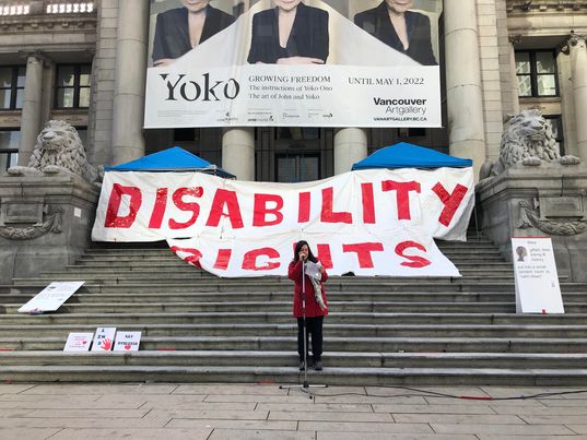 white woman with brown hair standing infront of a mic at the vancouver museum stairs are behind her with a large sign that says Disability Rights